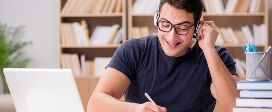 Man with glasses working from home on a computer and writing notes on a piece of paper.